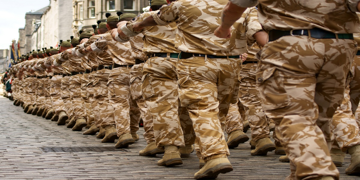 Photo taken from behind a troop of marching British soldiers in camouflage uniforms.