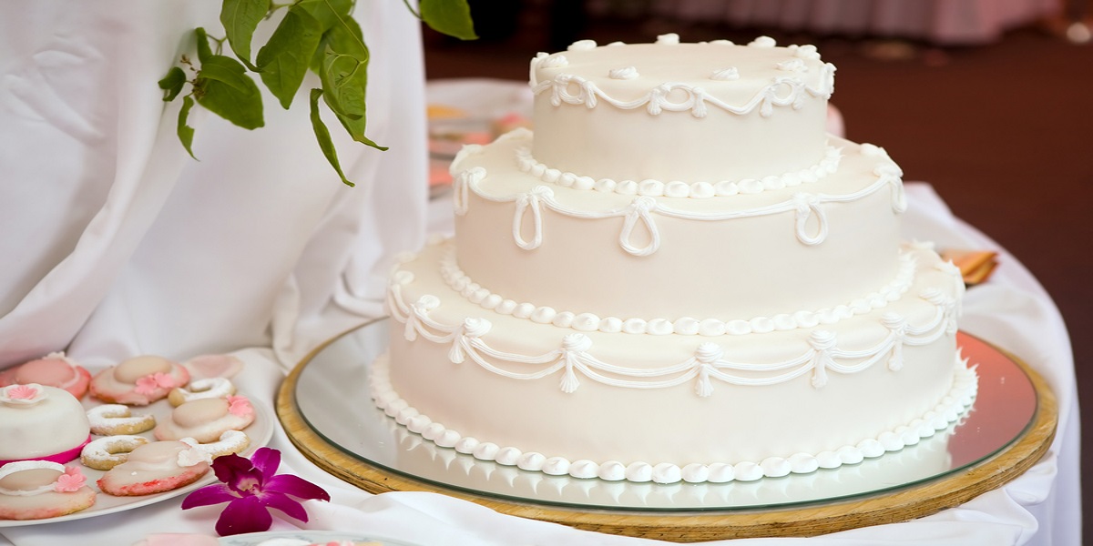 Three tier white wedding cake with pretty white piping decoration. Sitting on a decorated table beside a plate of wedding themed biscuits.