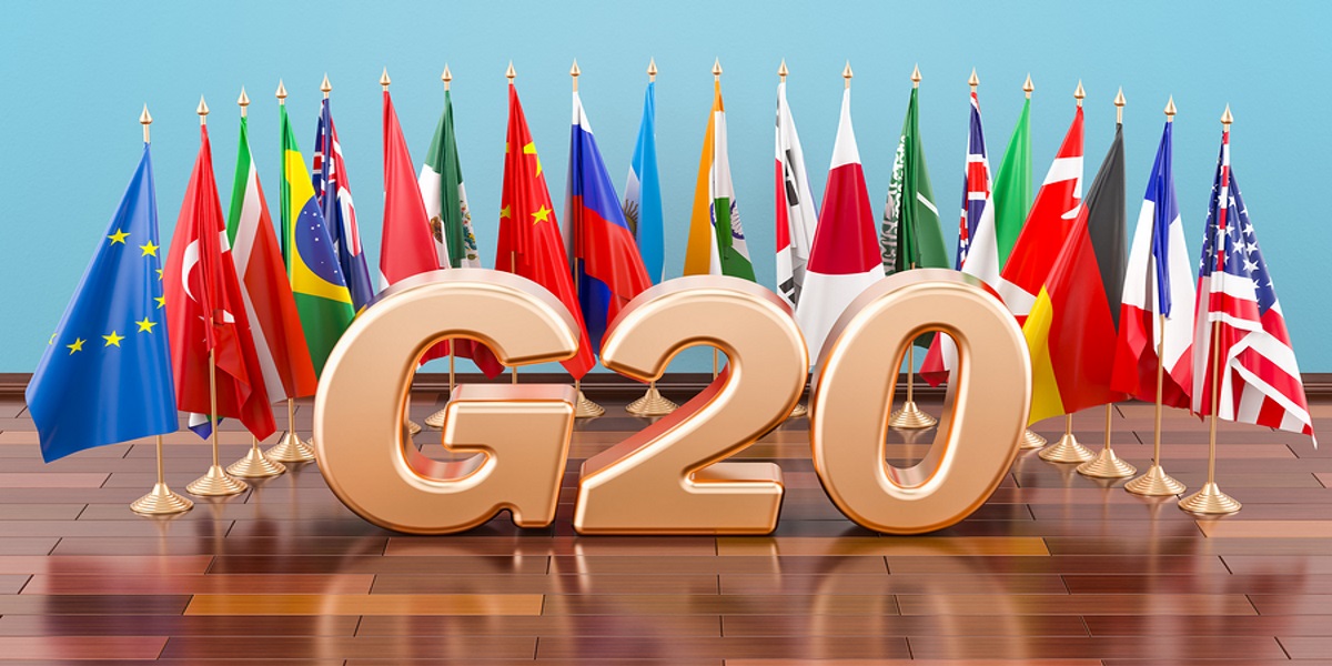 Rose gold G20 figures sitting in the centre of an arc of flags representing all 19 G20 countries, on a wooden table.