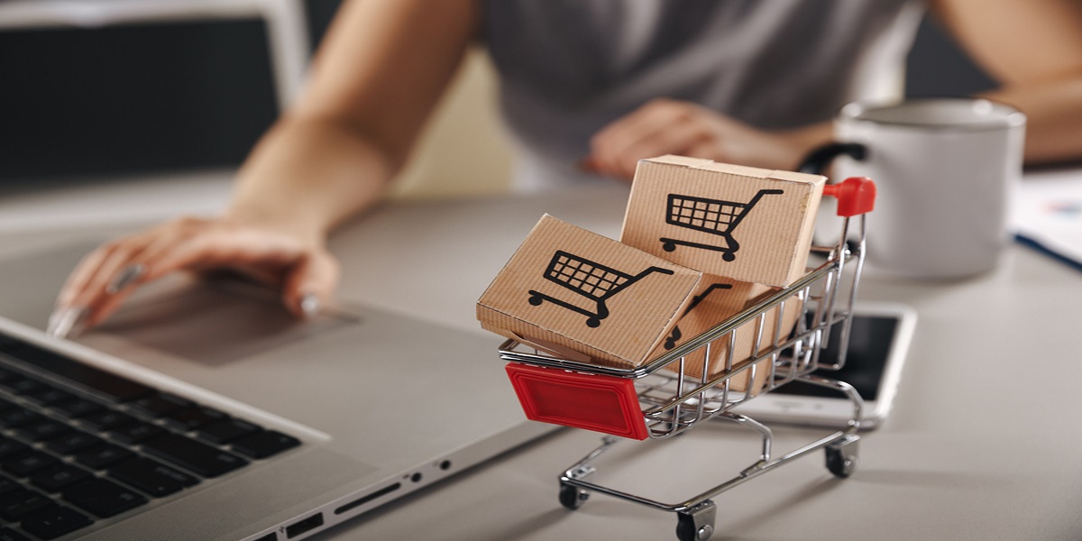 Foreground focus on a miniature shopping trolley containing mini boxes with common shopping cart icon on them. Representing ecommerce. Blurred background of a woman's hand over silver laptops' mouse, presumably ordering these items. Also mobile phone and mug on the desk.