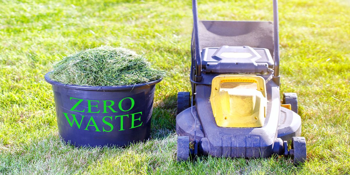 On grass, lawnmower on the right, bucket of newly mown clippings in a bucket labelled 'Zero Waste' on the left.