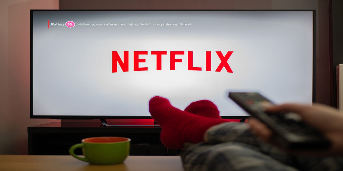 Netflix on large TV screen. Person's socked feet up on the table beside a cup, right hand pointing the remote control at the TV.
