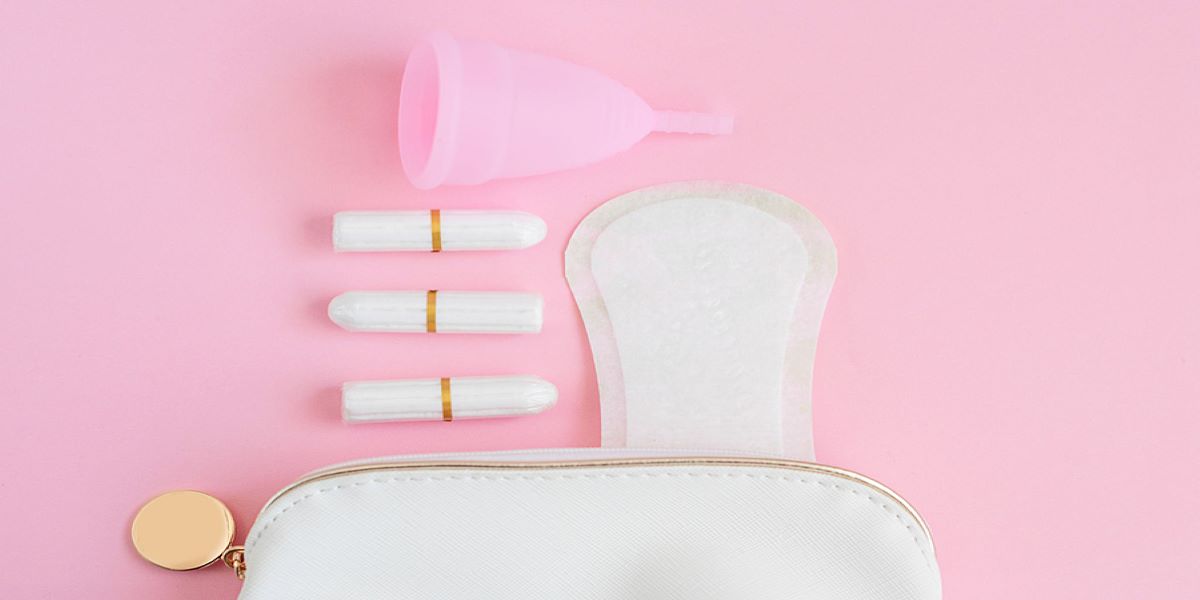 Pale pink background, with menstrual cup, 3 tampons, a sanitary towel and the top of a white bag.